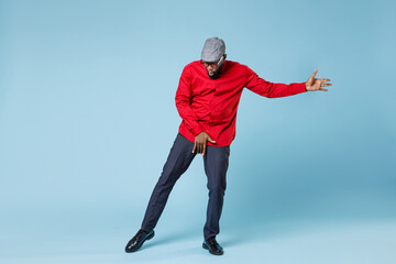 Full length of cheerful amazed young bearded african american man 20s wearing casual red shirt cap eyeglasses posing dancing put hand on crotch isolated on pastel blue wall background studio portrait.