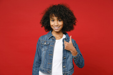 Smiling little african american kid girl 12-13 years old wearing casual denim jacket showing thumb up isolated on bright red color background children studio portrait. Childhood lifestyle concept. - obrazy, fototapety, plakaty