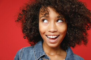 Close up of excited little african american kid girl 12-13 years old in denim jacket keeping mouth open looking aside isolated on red background children studio portrait. Childhood lifestyle concept.