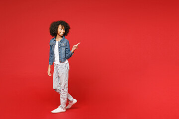 Fototapeta na wymiar Full length side view of smiling little african american kid girl 12-13 years old in denim jacket pointing index finger aside isolated on red background children portrait. Childhood lifestyle concept.