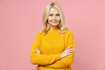 Smiling confident elderly gray-haired blonde woman lady 40s 50s years old in yellow casual sweater stand holding hands crossed looking camera isolated on pastel pink color background studio portrait.