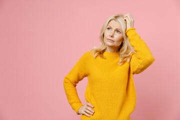 Worried pensive elderly gray-haired blonde woman lady 40s 50s years old wearing yellow casual sweater standing put hand on head looking aside isolated on pastel pink color background studio portrait.