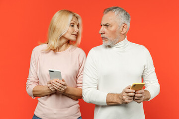 Displeased couple two friends elderly gray-haired man blonde woman wearing white pink clothes using mobile cell phone typing sms message isolated on bright orange color background studio portrait.