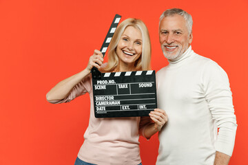 Cheerful couple two friends elderly gray-haired man blonde woman in white pink casual clothes hold classic black film making clapperboard isolated on bright orange color background studio portrait.