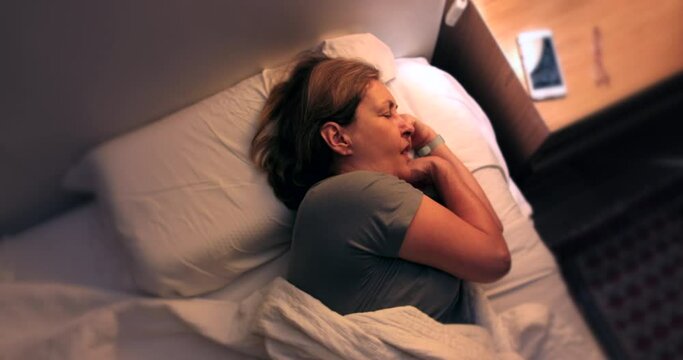 Tired Woman waking up in the middle of the night unable to sleep, turns on nightstand bed light and picks up phone