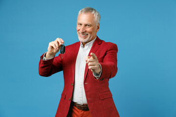 Cheerful elderly gray-haired mustache bearded business man wearing red jacket suit standing hold in hand car keys pointing index finger on camera isolated on blue color background studio portrait.