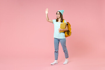 Full length of funny woman student in t-shirt hat glasses backpack hold notebooks waving greeting with hand as notices someone isolated on pink background. Education in high school university concept.