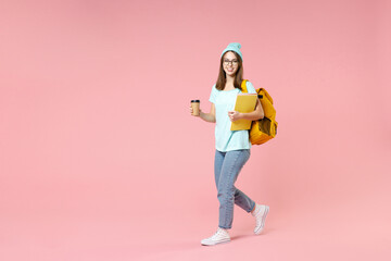 Full length side view of pretty young woman student in t-shirt hat glasses backpack hold notebooks paper cup of coffee or tea isolated on pink background. Education in high school university concept.