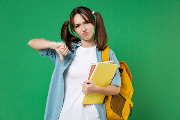 Displeased confused young woman student in blue shirt backpack hold notebooks showing thumb down isolated on green background studio portrait. Education in high school university college concept.