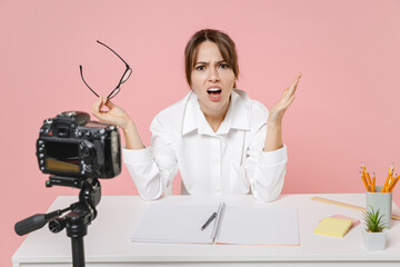 Angry irritated young woman tutor teacher in shirt glasses sit work at desk swearing conducting online lesson seminar recording video on camera isolated on pink background. Distance education concept.