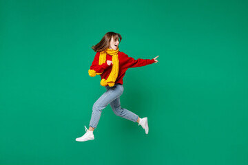 Fototapeta na wymiar Full length side view excited young brunette woman 20s in knitted red sweater yellow scarf jumping like running pointing index finger aside isolated on bright green color background studio portrait.