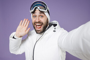 Close up of amazed skier man in windbreaker jacket ski goggles mask doing selfie shot on mobile phone greeting with hand spend weekend in mountains isolated on purple background. People hobby concept.