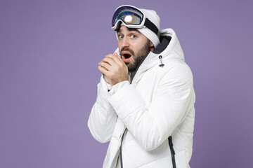 Freezing skier man in white windbreaker jacket ski goggles mask blowing on hands trying to keep warm spend weekend winter in mountains isolated on purple background. People lifestyle hobby concept.