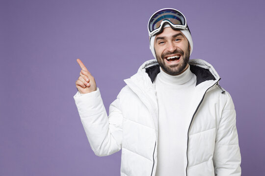 Cheerful skier man in white windbreaker jacket ski goggles mask pointing index finger up spend extreme weekend winter season in mountains isolated on purple background. People lifestyle hobby concept.