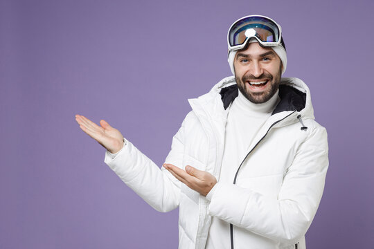 Cheerful skier man in white windbreaker jacket ski goggles mask pointing hands aside spend extreme weekend winter season in mountains isolated on purple background. People lifestyle hobby concept.