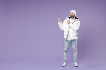 Full length of excited man in warm white padded windbreaker jacket hat pointing index fingers aside isolated on purple color background studio portrait. People lifestyle cold winter season concept. © ViDi Studio