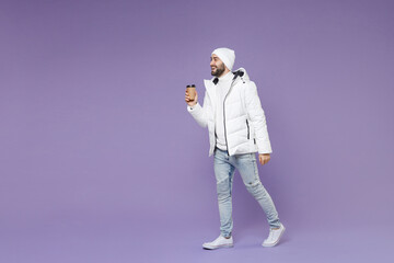 Full length side view of cheerful man in warm white windbreaker jacket hat hold paper cup coffee or tea isolated on purple background studio portrait. People lifestyle cold winter season concept. © ViDi Studio