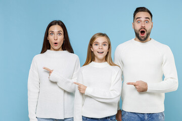 Shocked amazed young parents mom dad with child kid daughter teen girl in white sweaters point index fingers aside isolated on blue background studio portrait. Family day parenthood childhood concept.