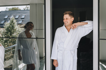 Obraz premium Stylish groom and sweet bride in white robes stand against the background of a mirrored wall and are reflected in an expensive hotel. Morning of the newlyweds. Wedding portrait.