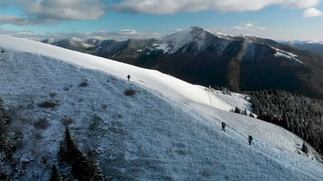 Aerial view of hiker friends walking on snow covered mountain ridge, ascending a summit in winter. 