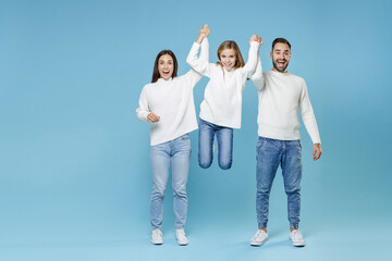 Full length of excited funny young happy parents mom dad holding child kid daughter teen girl in white sweaters jumping isolated on blue background studio portrait. Family day parenthood concept.