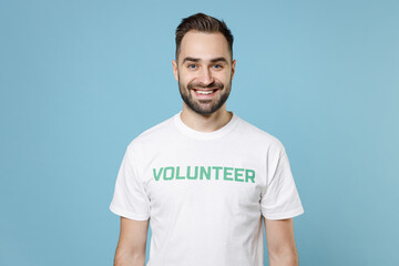 Fototapeta premium Smiling young bearded man 20s wearing white volunteer t-shirt standing looking camera isolated on blue color background studio portrait. Voluntary free work assistance help charity grace concept.