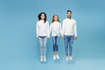 Obraz premium Full length of excited young parents mom dad with child kid daughter teen girl in white sweaters jumping holding hands isolated on blue wall background studio portrait. Family day parenthood concept.