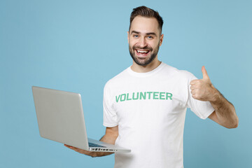 Cheerful young man in white volunteer t-shirt showing thumb up working laptop pc computer isolated...