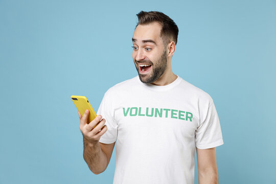 Excited young man in white volunteer t-shirt using mobile cell phone typing sms message isolated on blue color background studio portrait. Voluntary free work assistance help charity grace concept.