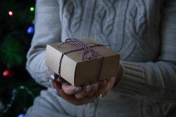 a girl in a beige knitted sweater holds a craft square gift box in her hands. Close-up