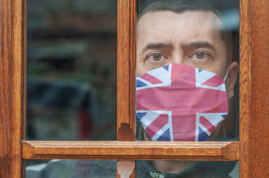 Young Man In A Protective Mask With A British Flag Stays At Home Looking Out The Window. Lockdown In The UK. Corona Virus Mutated Covid-19. Strengthening Quarantine. Coronavirus Is Mutating. Health