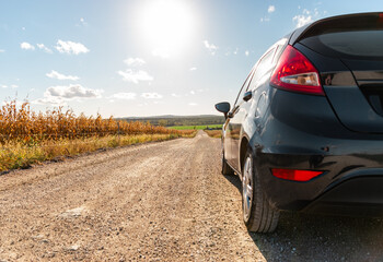 A car is standing on a pebbly road next to a corn field on sunny afternoon.