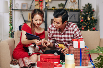 Positive Asian couple with small dog following tutorial on tablet computer when decorating...