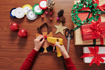 Hands of person glueing red nose on deer gift package at table with presents, ribbons and baubles