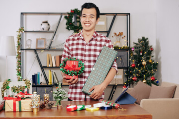 Fototapeta na wymiar Happy handsome young Asian man standing at table with wrapped presents and Christmas wreath in hands