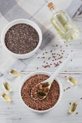 Brown flax seed or linseed and chia in small bowl and gelatin capsules with omega oil on a white wooden background.