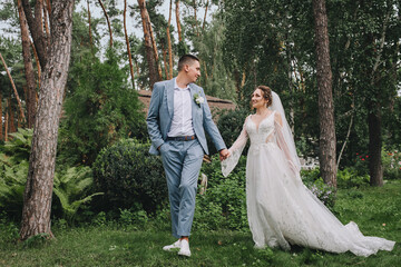 Young stylish groom in a blue suit and a beautiful bride in a white lace dress walk holding hands in the garden with green plants in nature with pine trees. Wedding portrait of the newlyweds.