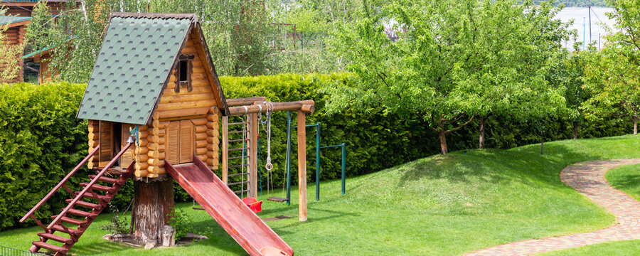 Small wood log playhouse hut with stairs ladder and wooden slide on children playground at park or house yard. Green grass lawn garden and paved pathway background on bright sunny day. Panoramic view