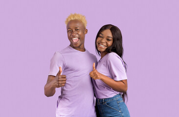 Happy black couple showing thumbs up gesture, approving something over lilac studio background