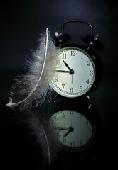 Black alarm clock on a mirror surface, feather on black background