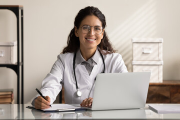 Portrait of confident young Caucasian female doctor in white medical uniform sit at desk working on...