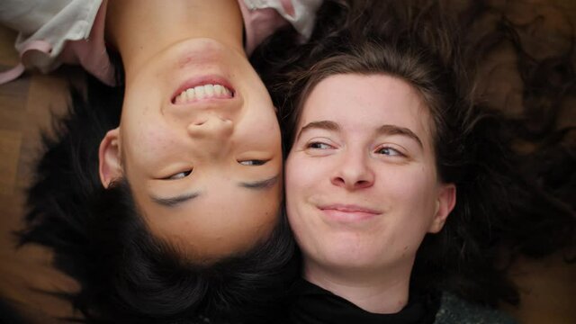 Two happy and funny women lying on floor, looking at each other and smiling. Portrait of Asian and Caucasian girls having fun and laughing. Interracial friendship or mixed-race lesbian couple. 4K.
