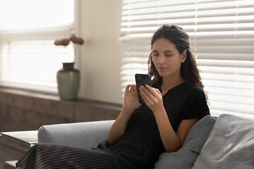 Young Caucasian woman sit relax on couch use modern cellphone gadget texting or messaging. Millennial female user or client look at smartphone screen, shopping online or browsing internet on device.