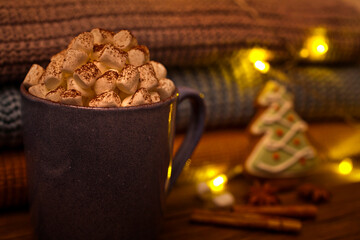 Cocoa drink with marshmallows in a blue ceramic cup, sweet spices, fir-shaped gingerbread cookie, a stack of knits and electric garland lights on a wooden background.