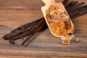 Brown sugar crystals in a wooden scoop with vanilla beans on a wooden background.