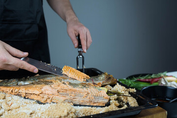Chief cook is breaking salt and carving fish baked in salt crust. Seafood cooking. Salmon or...