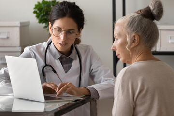 Young female doctor use computer consult mature patient in modern hospital. Woman GP and senior client look at laptop screen, discuss treatment or therapy in clinic. Elderly healthcare concept.