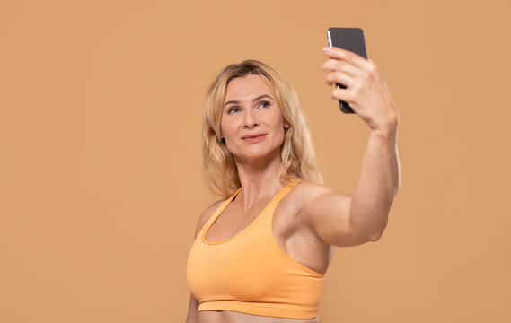 Selfie or stock video of coach. Adult smiling lady trainer in sports bra holds smartphone and makes photo