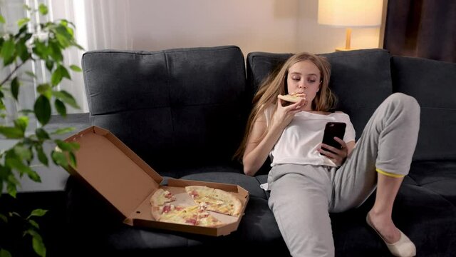 Cheereful european young woman extremely frustrated. Bored girl sitting on sofa at modern living room and eating tasty pizza. Millennial female using smartphone