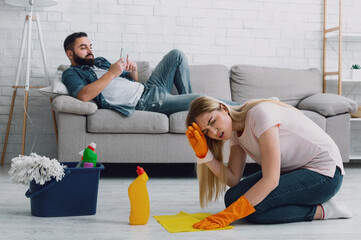 Woman suffers from headache and does cleaning at home, man with beard lies on couch and plays on...